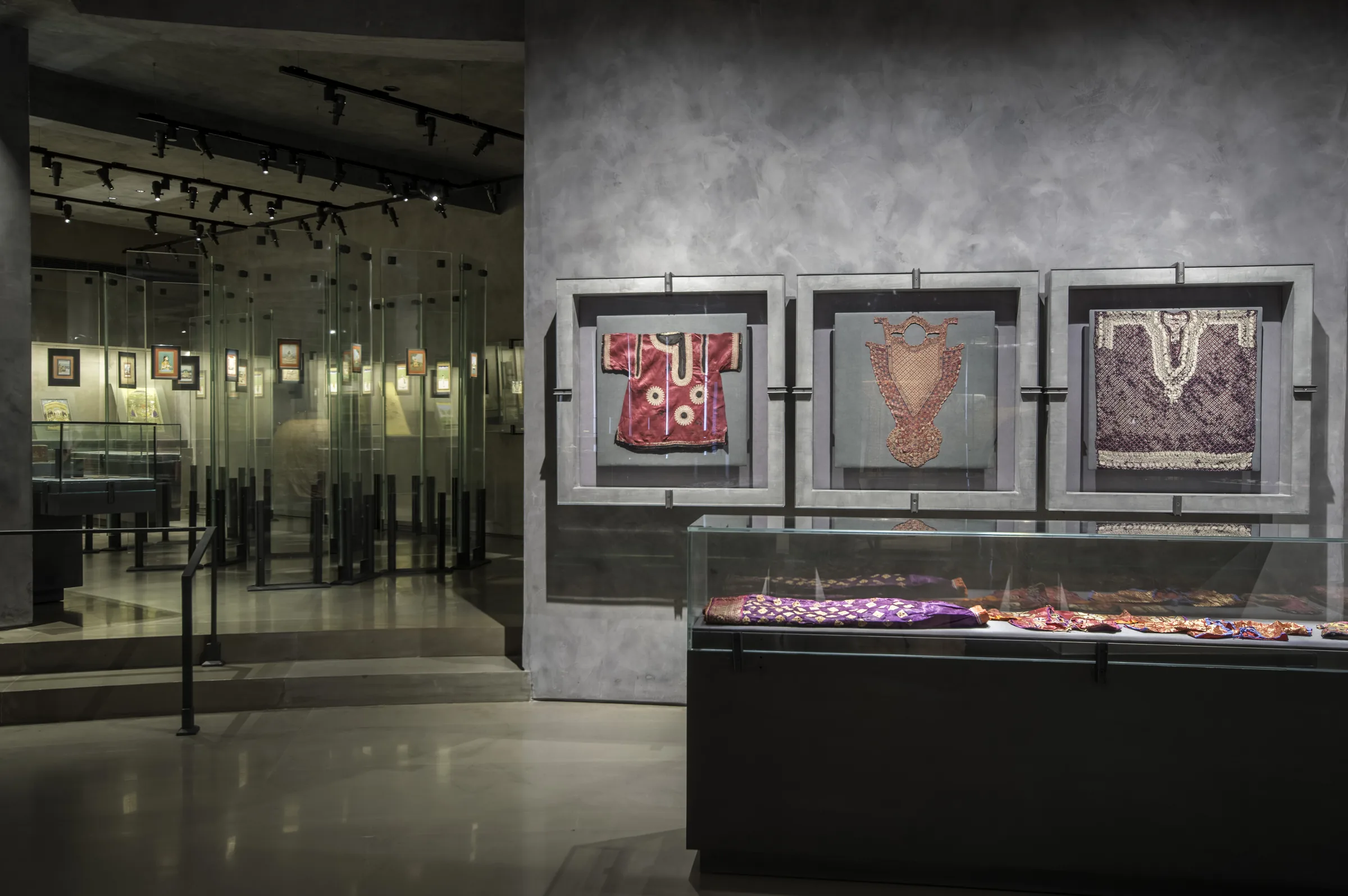 Three framed garments on a stone-grey wall, with a case of embroidered fabrics beneath.